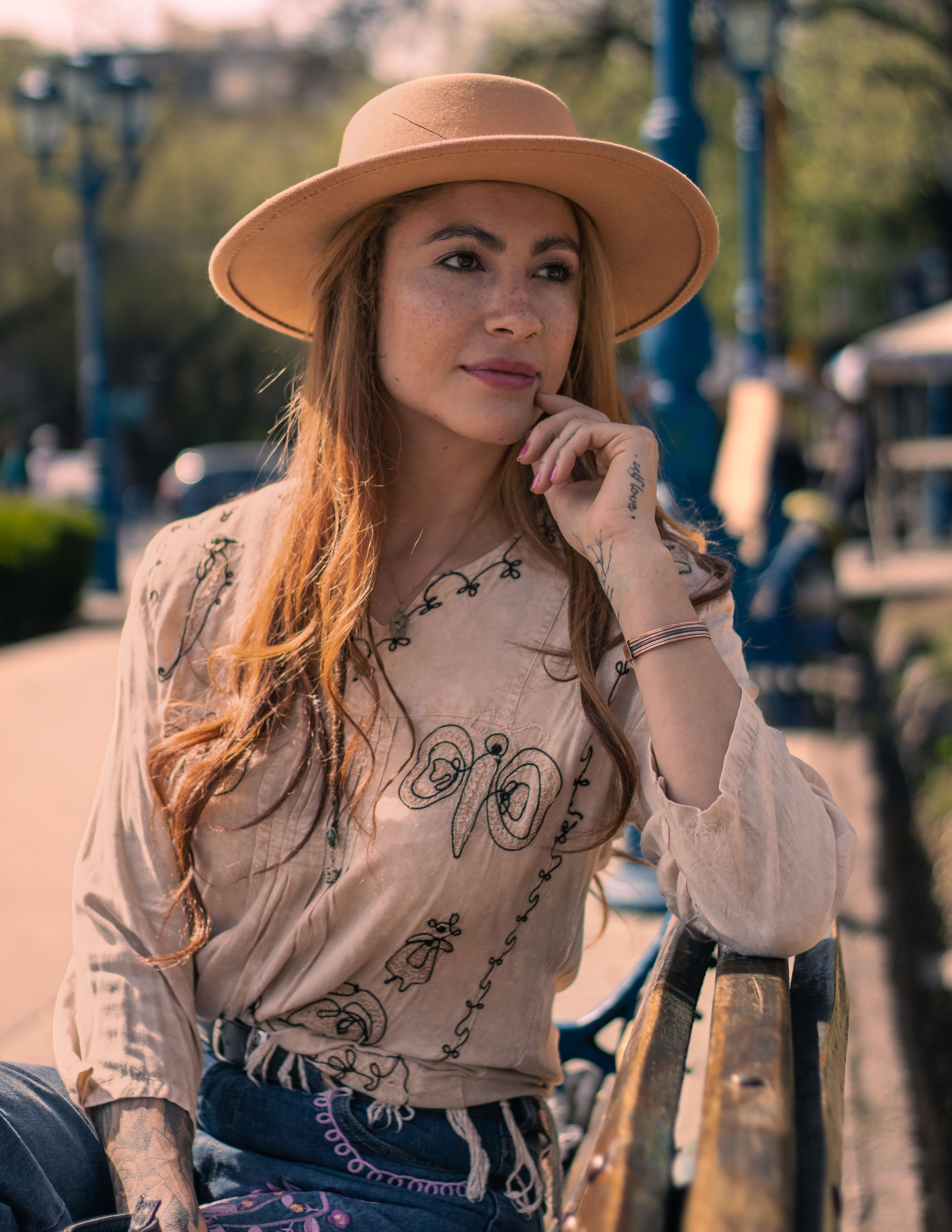 Mulher com um chapéu chique e blusa bordada senta-se em um banco de parque desfrutando um dia ensolarado.
