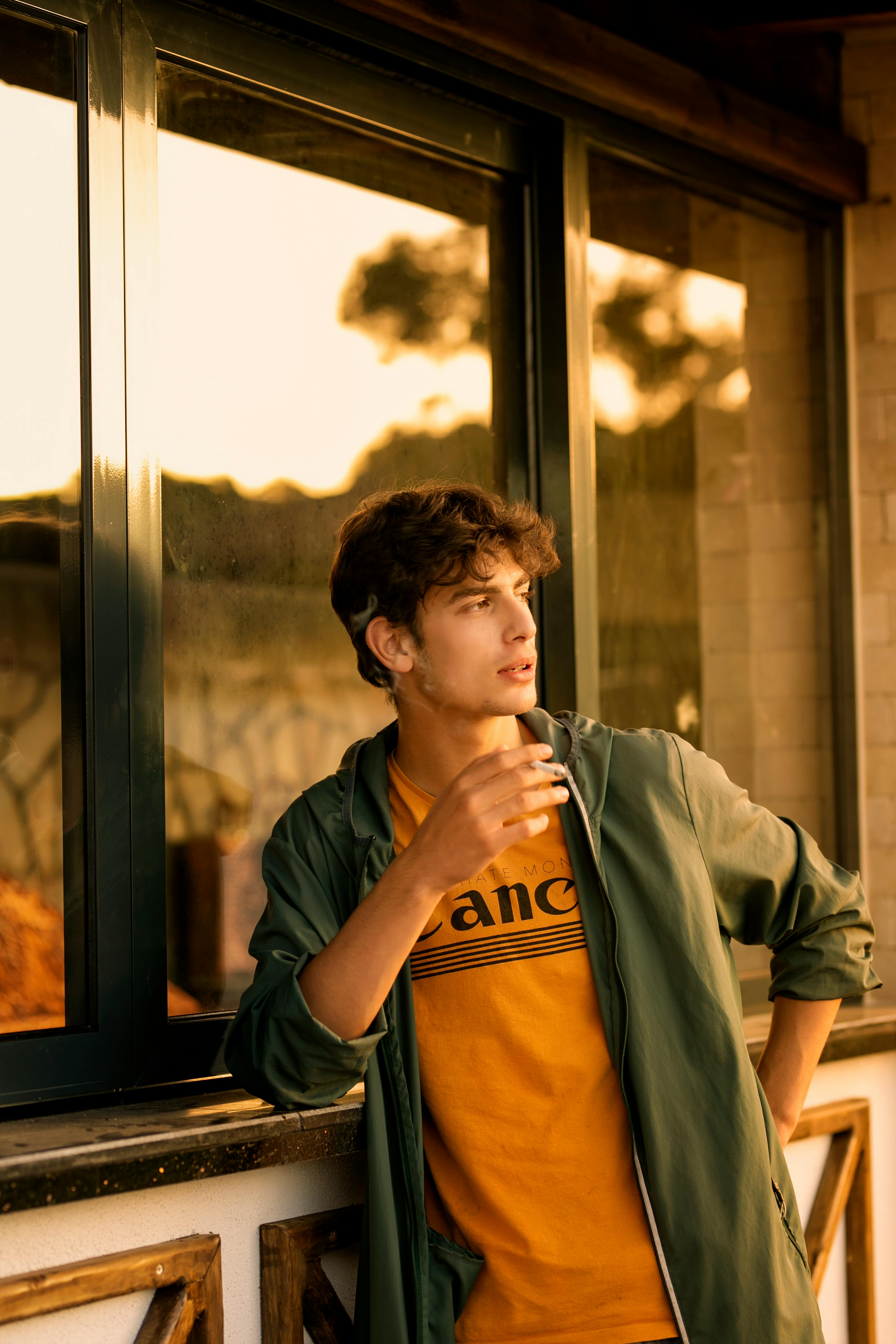 A young man in a yellow shirt and green jacket gazes thoughtfully out of a window at sunset.