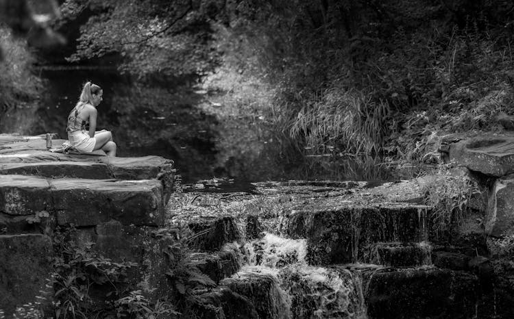 Grayscale Photo Of A Woman Sitting On Rock Near Waterfalls