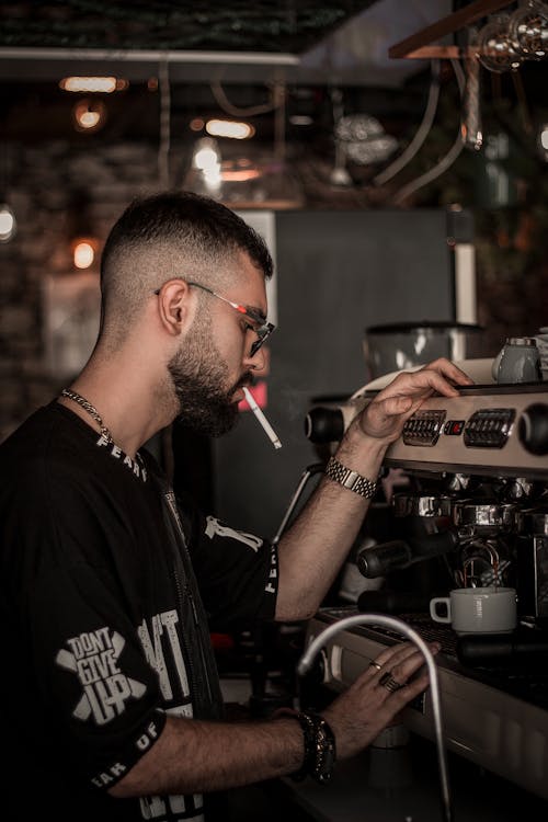 A Man in Black Shirt Smoking Cigarette while Making Coffee