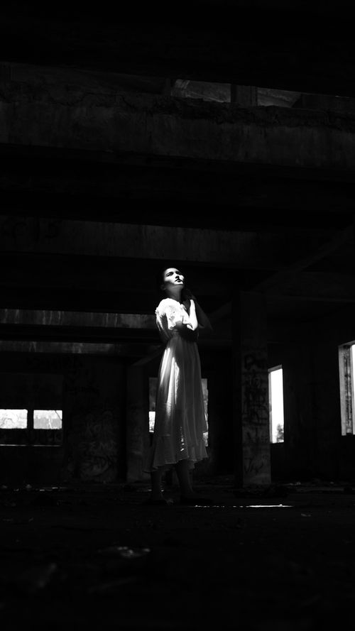 Free Dramatic black and white photo of woman in a dress standing in a dimly lit abandoned building. Stock Photo