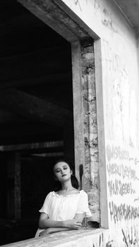 Black and white portrait of a woman by a graffiti-covered window in an abandoned building.