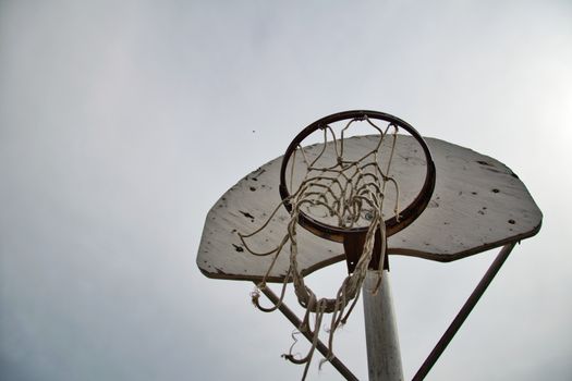 Low angle view of an old basketball hoop with worn net under a cloudy sky, emphasizing outdoor sports nostalgia.