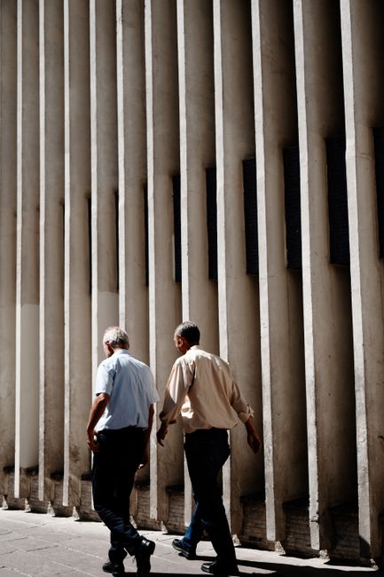 Two men walking beside a textured urban wall on a sunny day, creating a vintage feel.