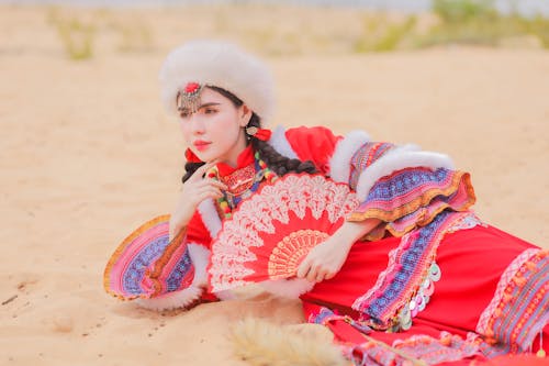 Woman in Red Traditional Dress Holding Hand Fan while Lying on Sandy Ground