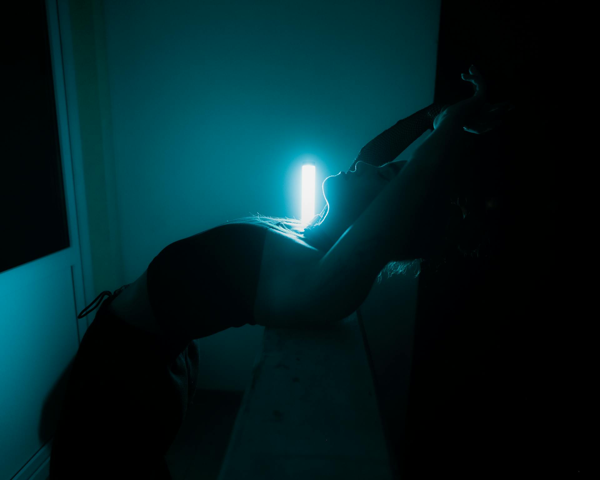 A dramatic silhouette of a woman leaning against a glowing tube light in a dark room.