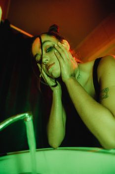 A woman washes her face under a flowing faucet, surrounded by dramatic lighting. Indoor shot.