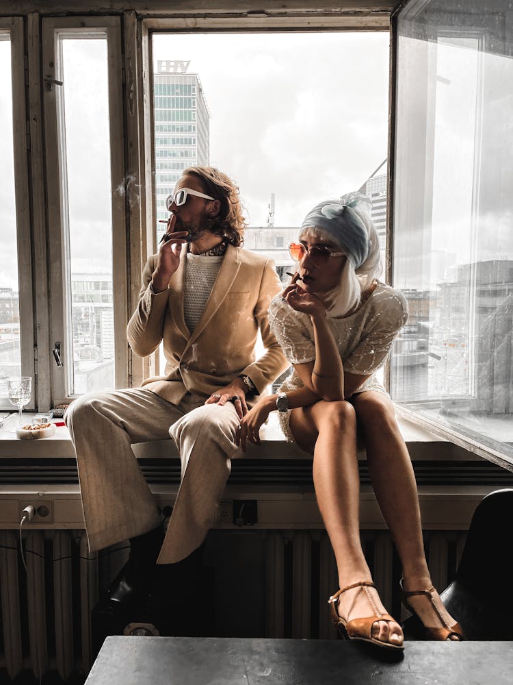 Man And Woman Sitting On A Windowsill Of A High Rise Building And Smoking Cigarettes 