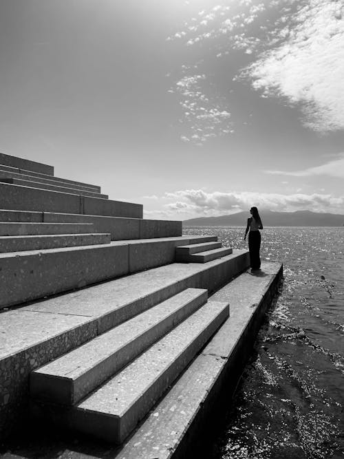 Grayscale Photo of Woman Standing on Concrete Stairs Near Body of Water