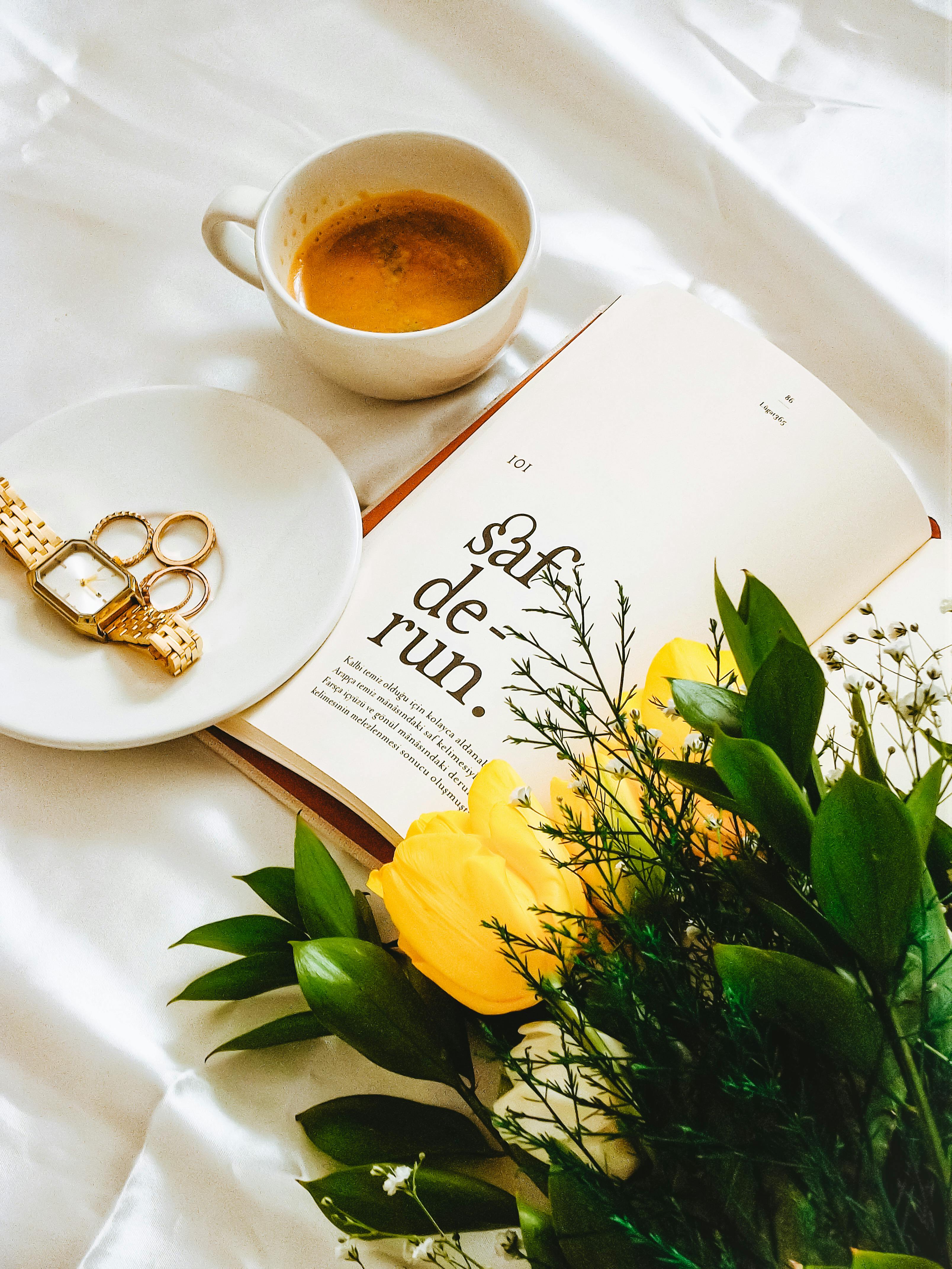 A cozy still life with coffee, book, flowers, jewelry, and wristwatch on a white surface.