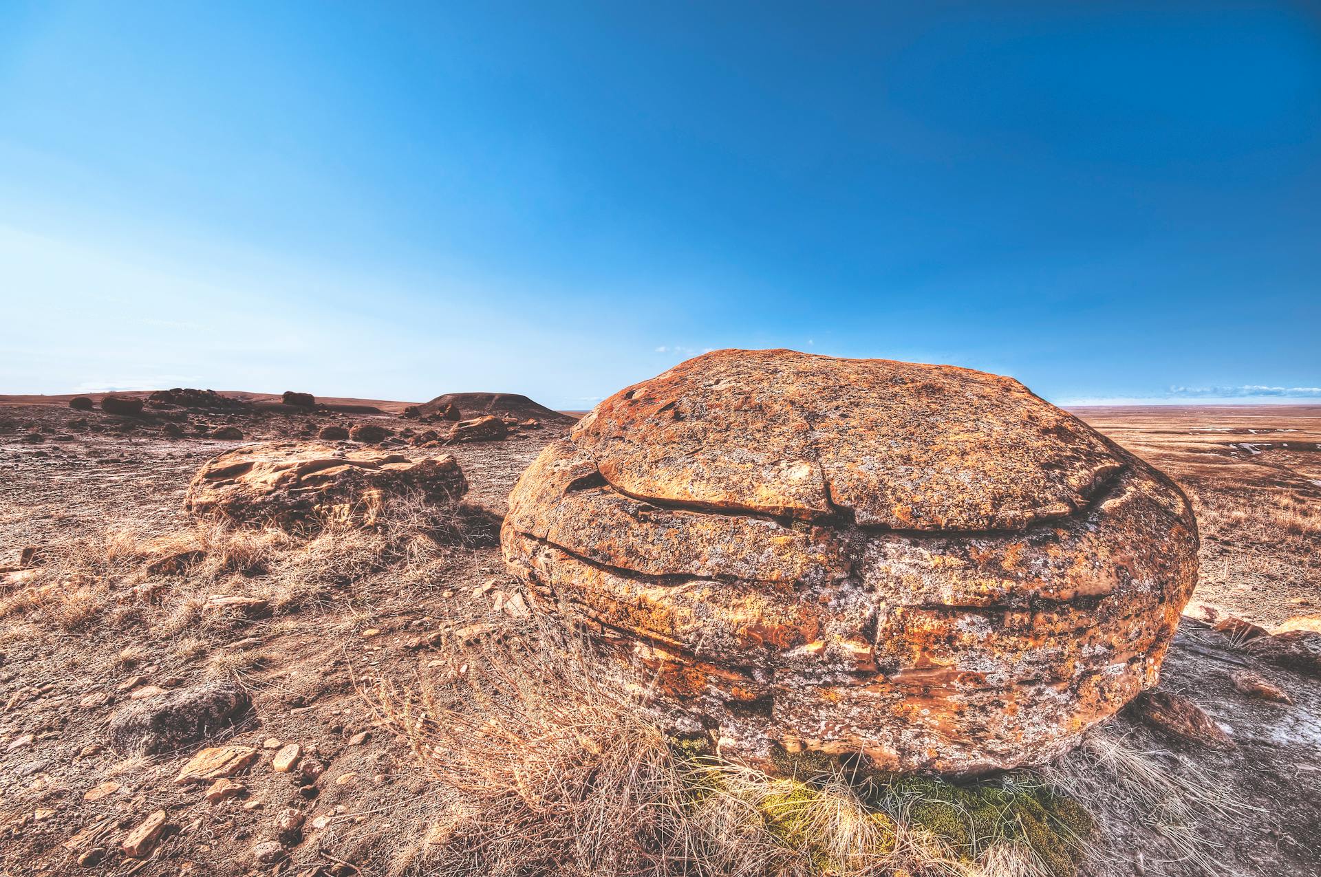 Expansive arid desert landscape featuring a prominent sandstone boulder under a clear blue sky.