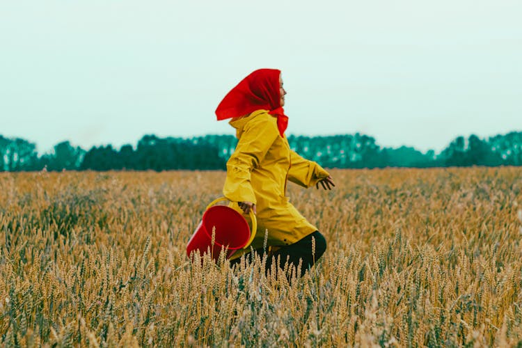 Woman In Yellow Coat And Handkerchief Running In Field With Bucket