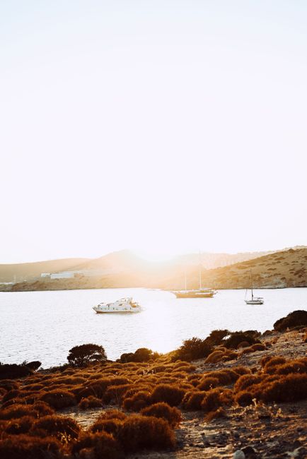 Tranquil scene of boats on a calm sea at sunset, with a rugged coastline in the foreground.