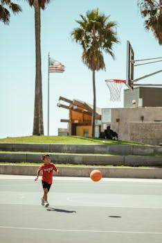 Child enjoying a game of basketball on an outdoor court with palm trees and a blue sky backdrop.