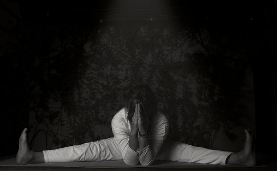 A woman practicing a yoga pose with a leg split indoors in monochrome lighting.
