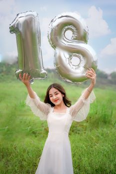 Cheerful young woman in a white dress holding '18' balloons on a sunny day in a grassy field.