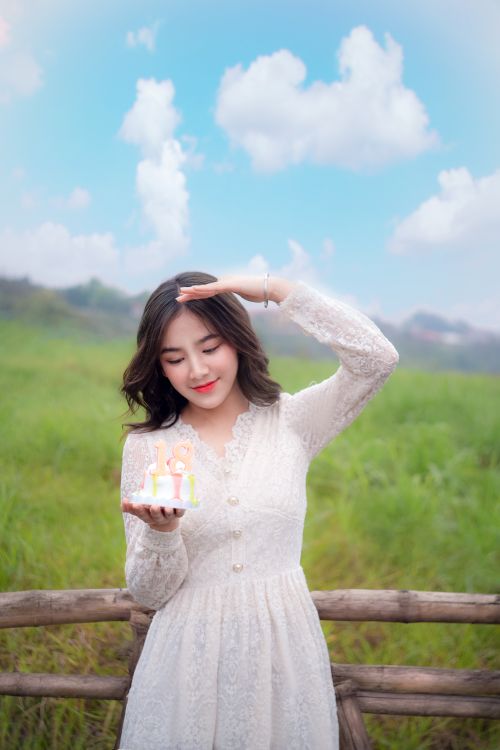 Free A young woman in a white dress enjoys a sunny day outdoors, holding a small cake. Stock Photo