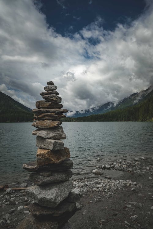 Stack of Stones on Shore Near Mountain Under Cloudy Sky