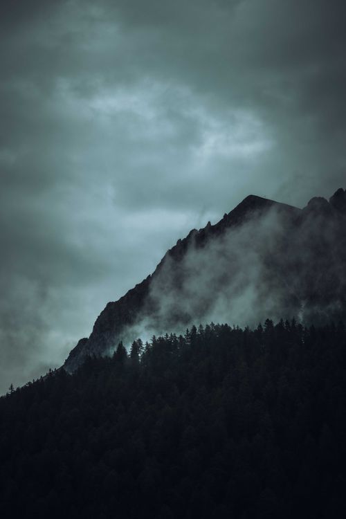 Free Dramatic mist envelops a rugged mountain peak above a dark forest, creating a moody landscape. Stock Photo