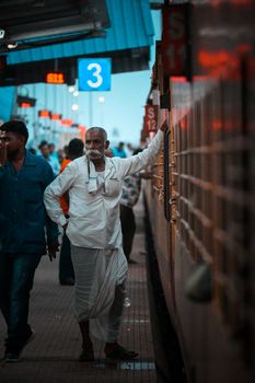 An elderly man in traditional attire stands on a railway platform beside a train.