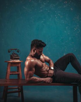 Strong shirtless man with beard posing on a bench next to vintage telephone indoors.