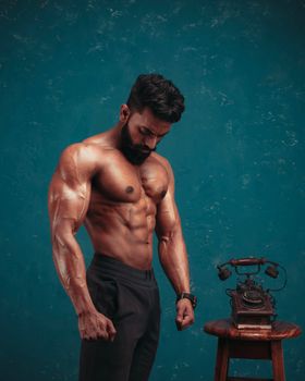 Portrait of a muscular man standing shirtless with a vintage telephone on a table.