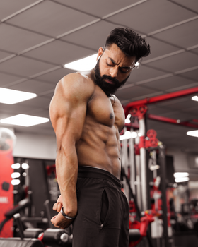 Shirtless muscular man flexing his arms in an indoor gym setting, showcasing fitness and strength.