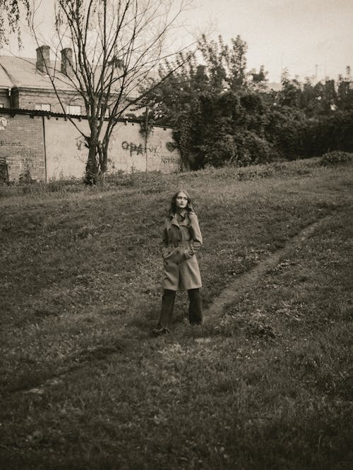 Grayscale Photo of a Woman in a Coat Walking on a Grassy Field