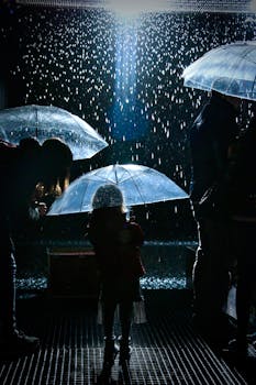 Silhouetted figures holding umbrellas in a nighttime rain shower, creating a dramatic and moody scene.