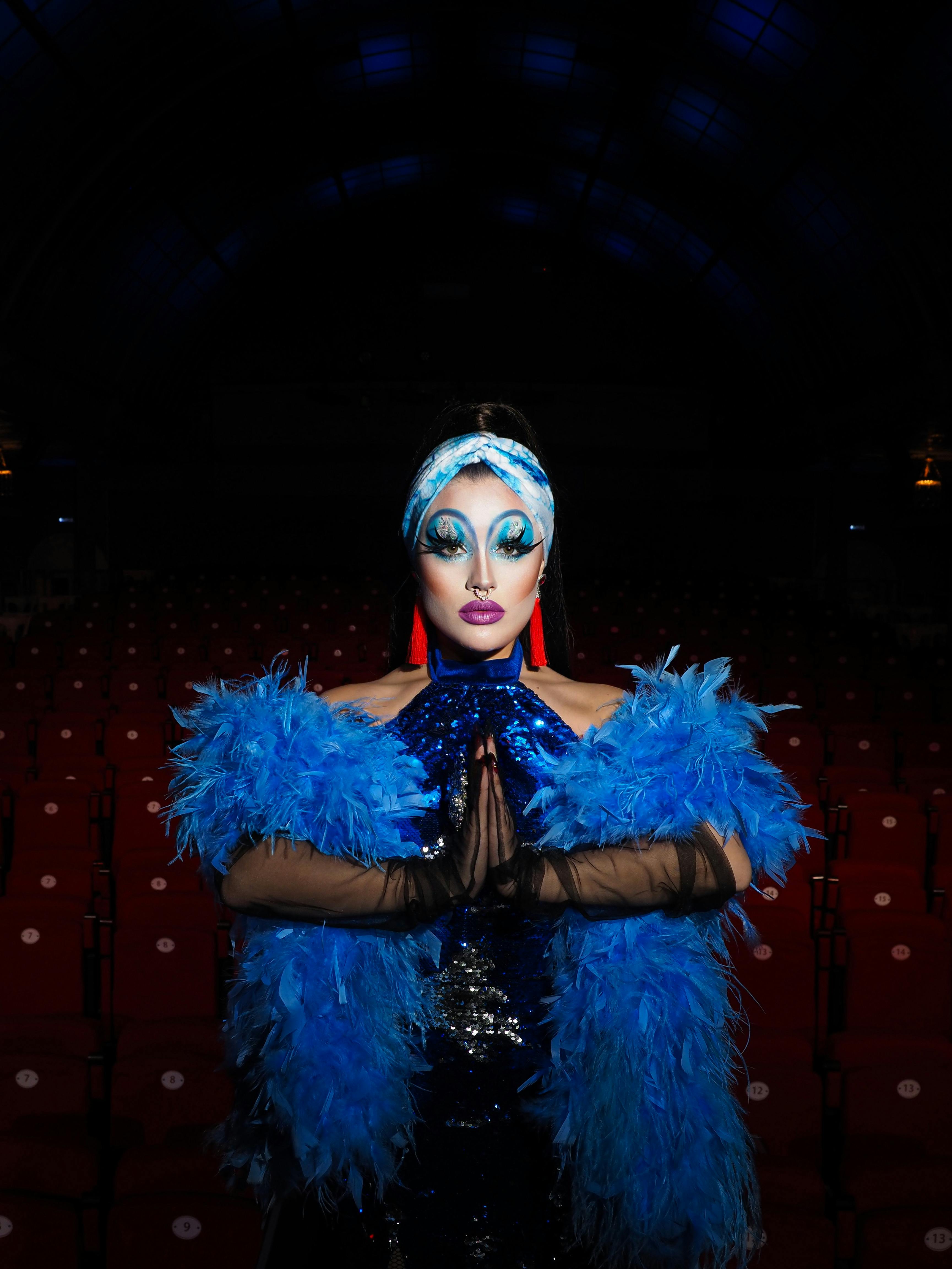 Free Performer in blue feather boa and bold makeup on a dimly lit stage, conveying glamour and theatricality. Stock Photo