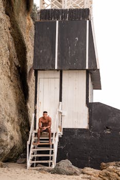 A man sits on stairs beside a rustic beach structure by a rocky cliff, embodying relaxation and summer vibes.
