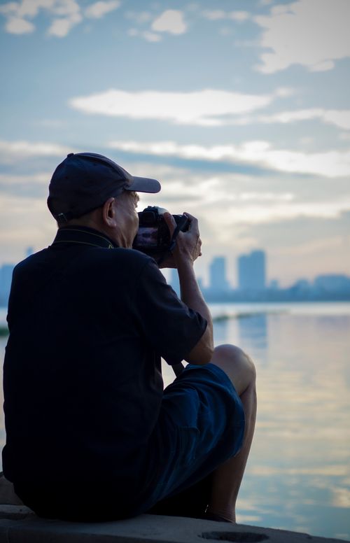 Free A senior man sitting and photographing a waterfront at sunset with a DSLR camera. Stock Photo