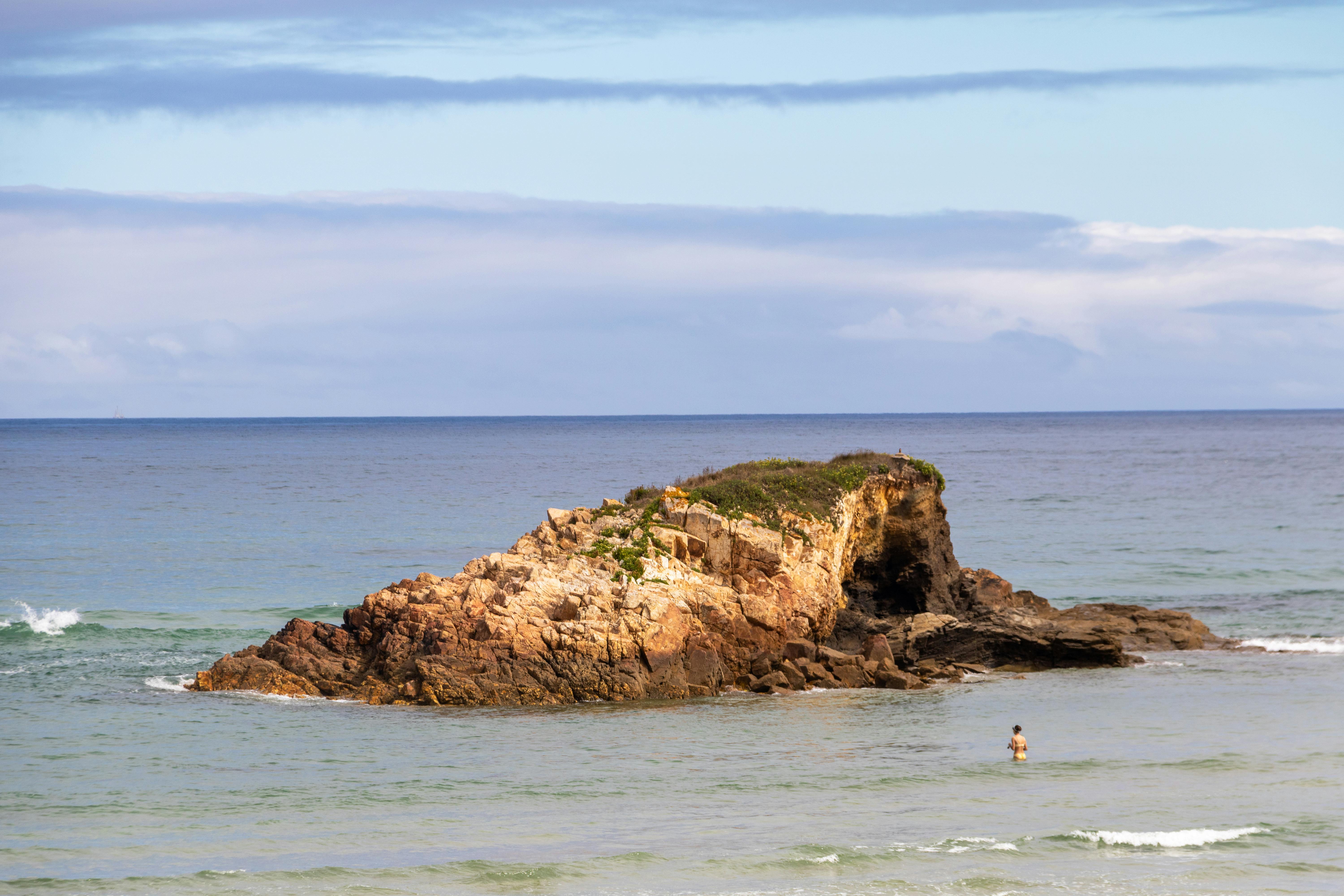 Rock formation on a beach in Lugo, Spain with a lone swimmer and clear skies.