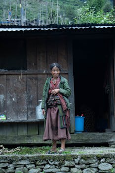 An elderly woman stands in front of a rustic wooden cottage, showcasing traditional attire in a rural setting.