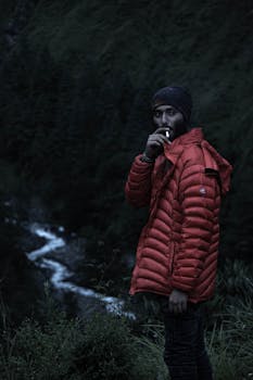 A man wearing a red bubble jacket and beanie, smoking outdoors by a mountain stream.