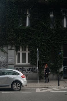 A city sidewalk with a building covered in climbing vines and a parked car. Two people walking by.