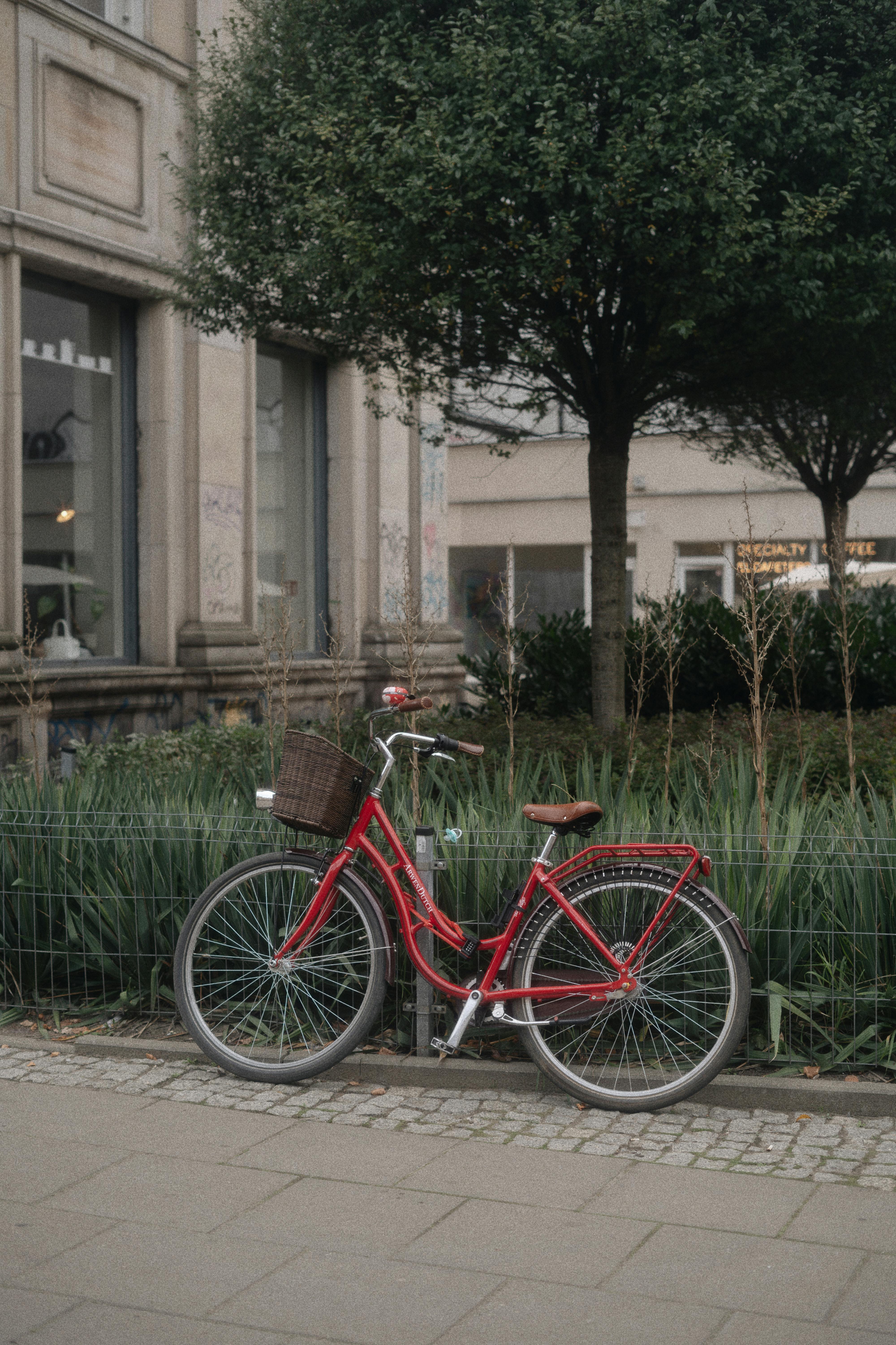 A red bicycle with a basket parked by a lush urban garden on a sidewalk.