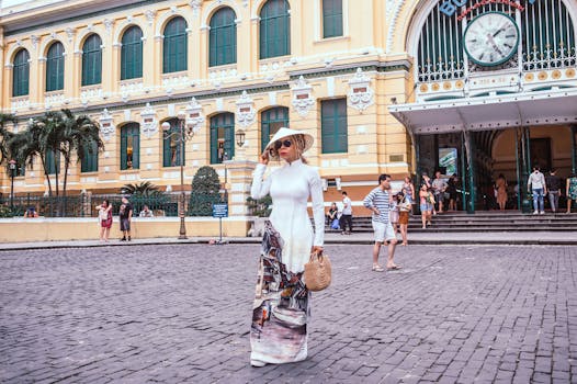 Elegant woman in ao dai and hat poses at the historic Saigon Central Post Office in Vietnam.