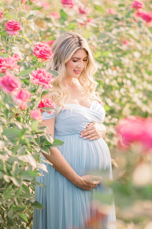 A Woman in Blue Dress Touching Her Baby Bump while Standing on a Flower Field