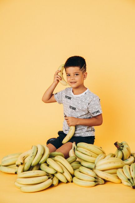 Young boy in printed shirt posing with bananas on a yellow background in a studio.