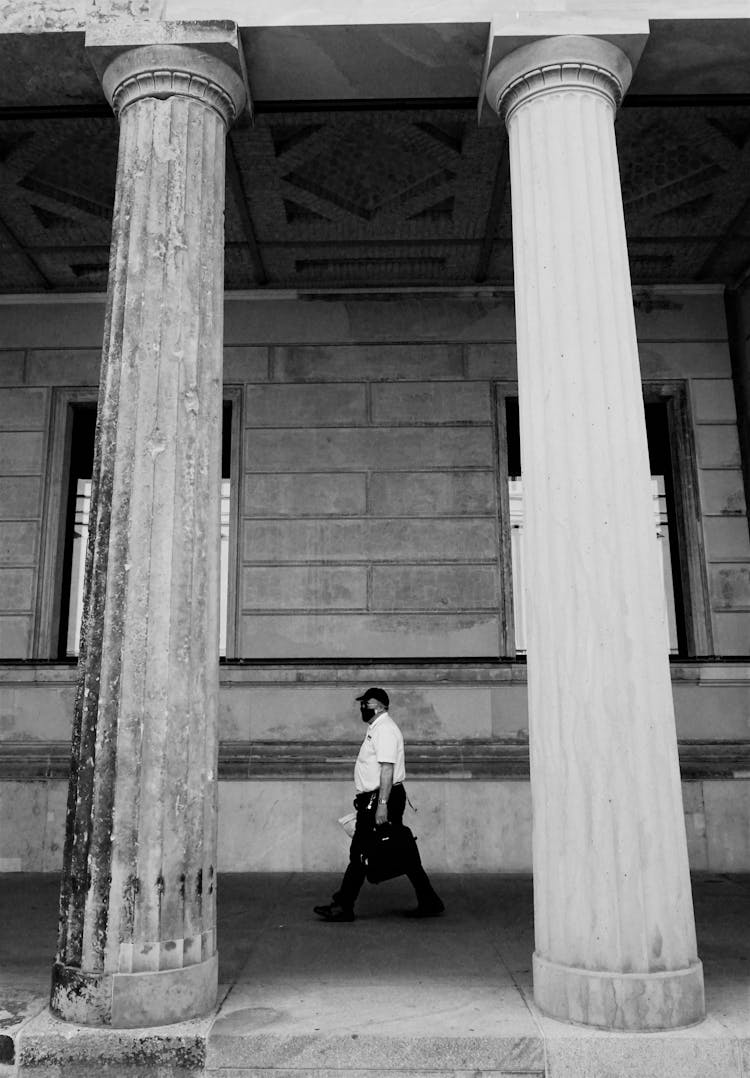 Man With Face Mask Walking On Concrete Pavement Between Pillars