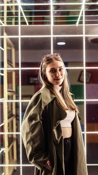 Young woman in a fashionable coat posing confidently against a neon-lit grid backdrop.