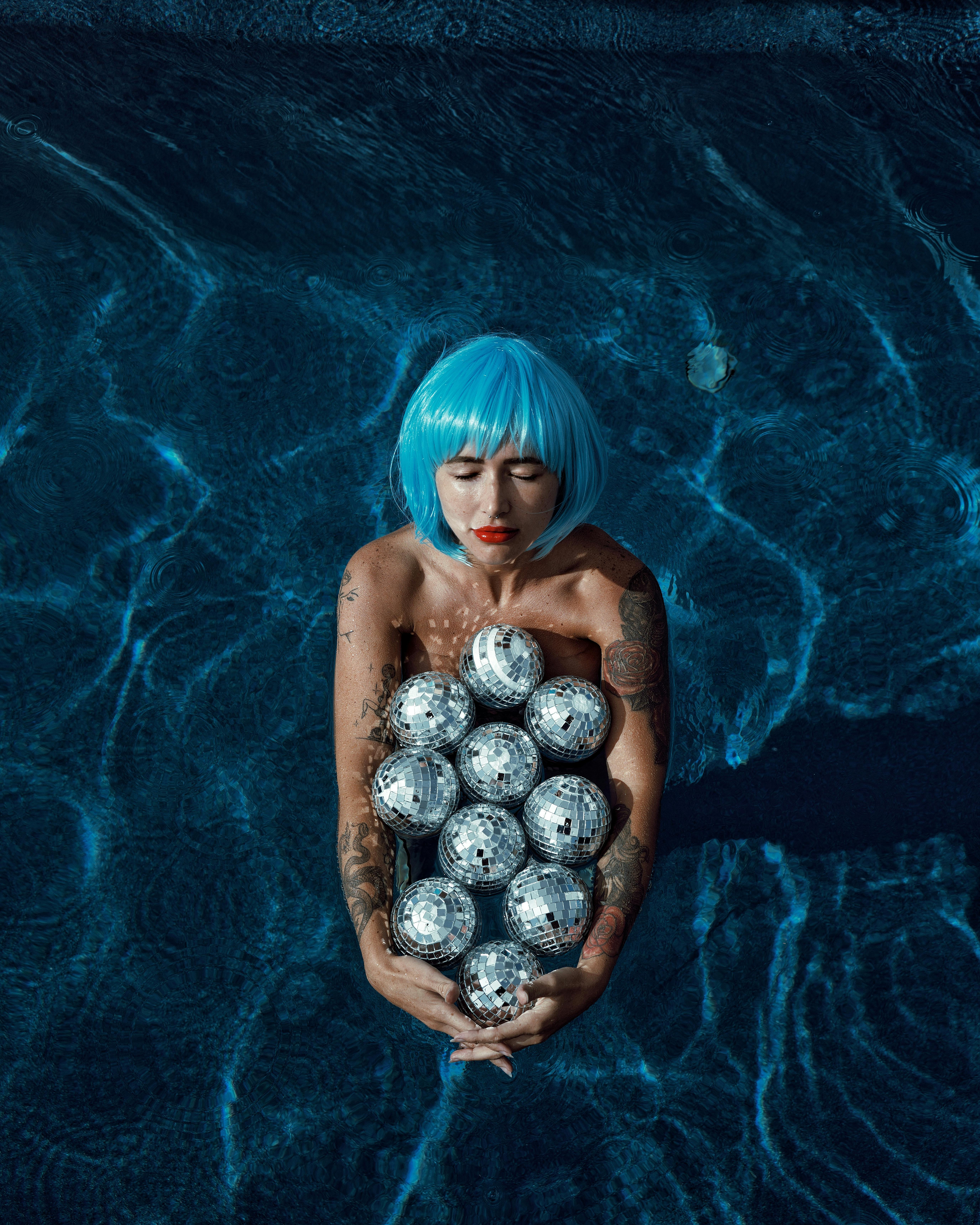 Creative shot of a woman in a blue wig, holding disco balls in a swimming pool.