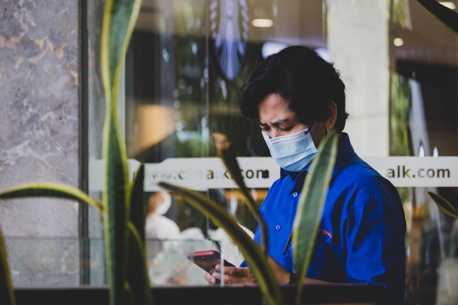 Persona con máscara facial y camisa azul usando smartphone en interiores.