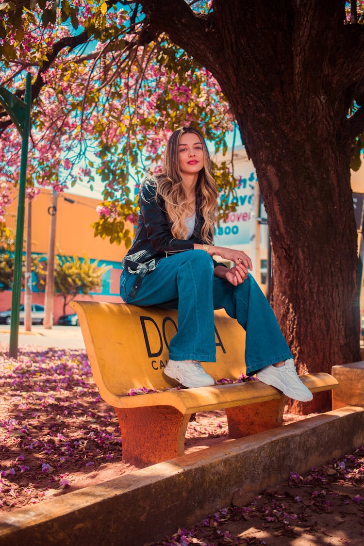 A Woman In Black Jacket Sitting On A Bench Near Big Tree While Looking At The Camera