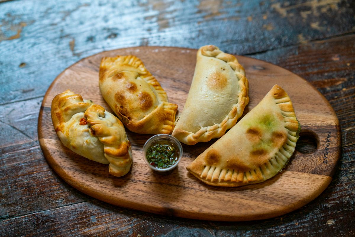 Four baked crimped empanadas on a wooden board with a small cup of chimichurri