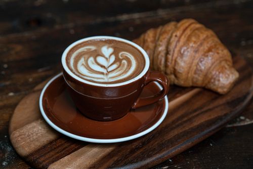 Free A close-up of a latte with beautiful art served alongside a croissant on a rustic wooden board. Stock Photo