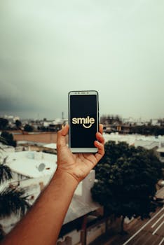 Person holding a smartphone with 'smile' text on screen against an outdoor urban backdrop in India.