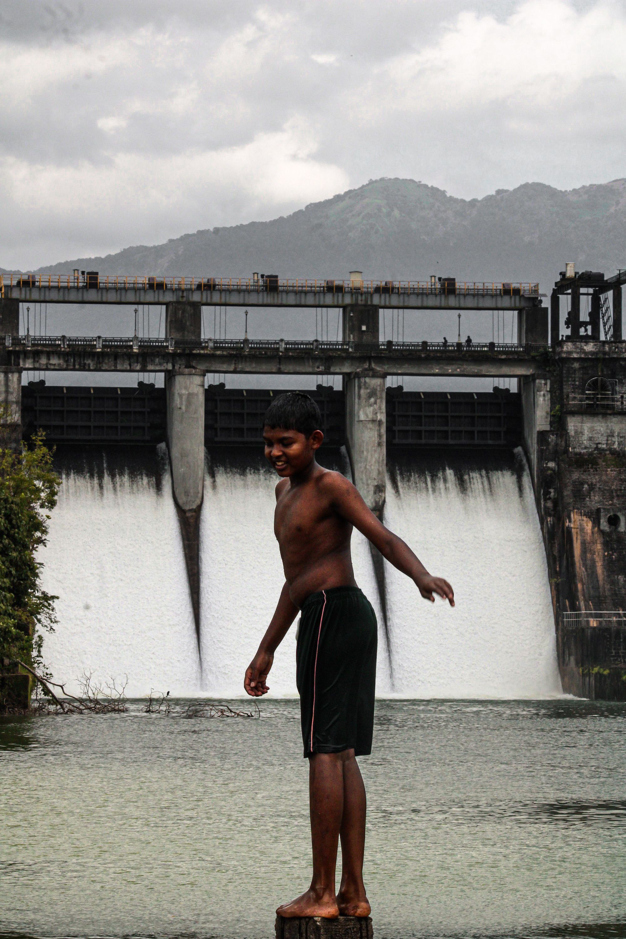 A boy stands confidently near a large dam with cascading water.