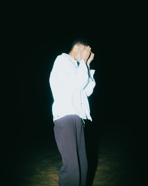 Man covers face in dramatic lighting, standing isolated in nighttime scene.